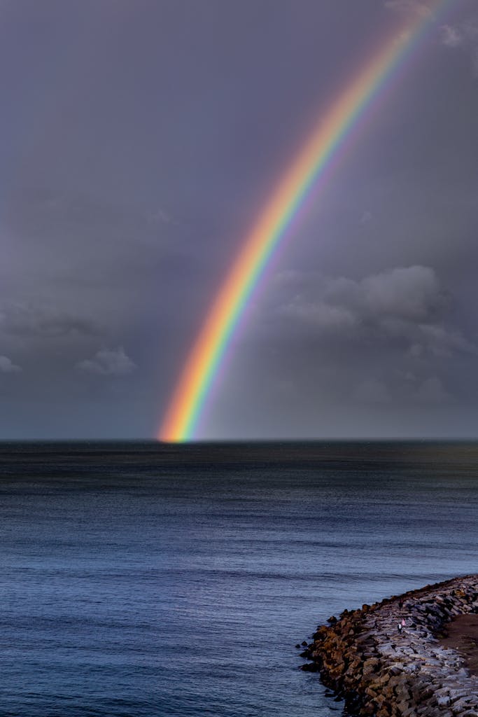 Stunning rainbow arches over deep blue ocean with rocky shoreline under cloudy sky, creating a serene seascape.