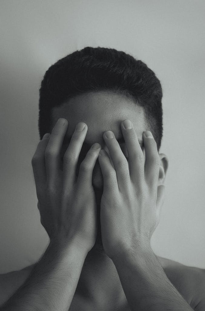 A black and white portrait of a young man covering his face with hands, depicting anxiety and stress.